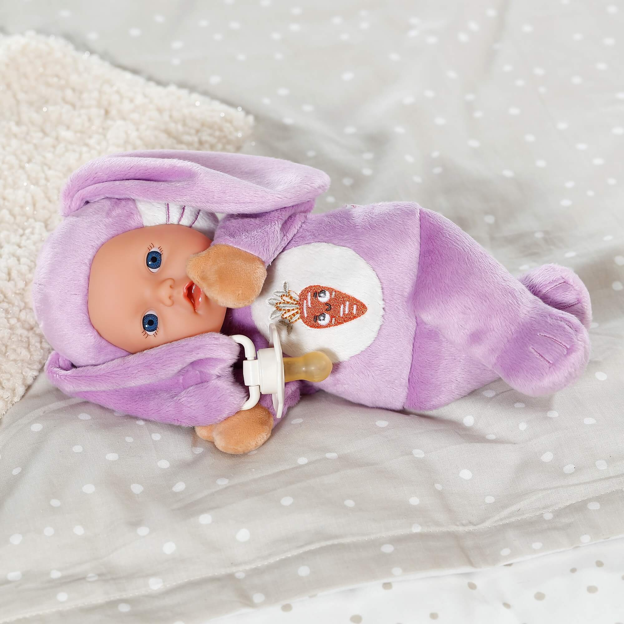 Muñeca bebé en traje de conejito morado con parche de zanahoria y chupete sobre tela de lunares.