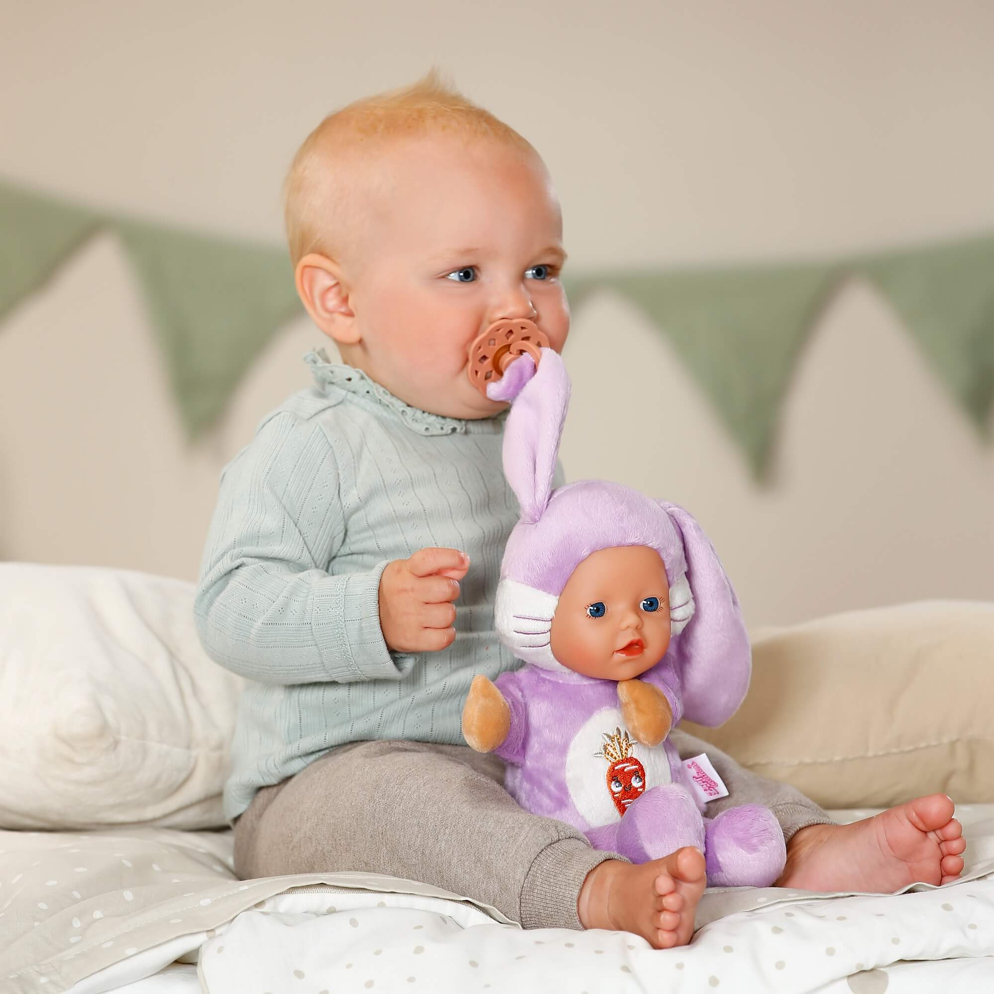 Un bebé con chupete sostiene un muñeco de conejito morado, sentado en una cama.