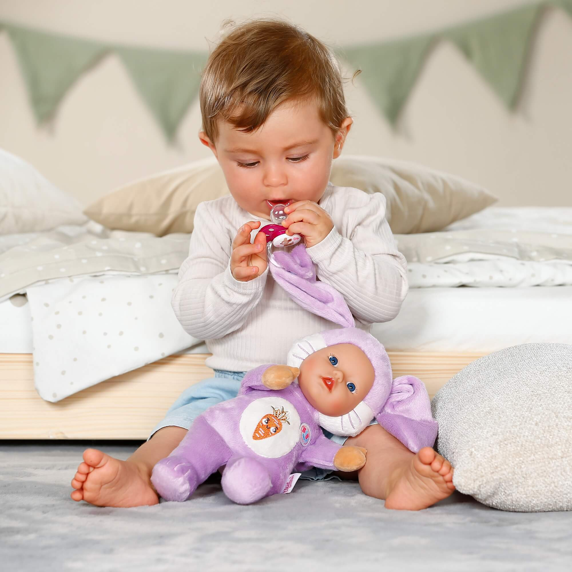 Un bebé se sienta en el suelo jugando con una muñeca de conejito morada y un chupete.