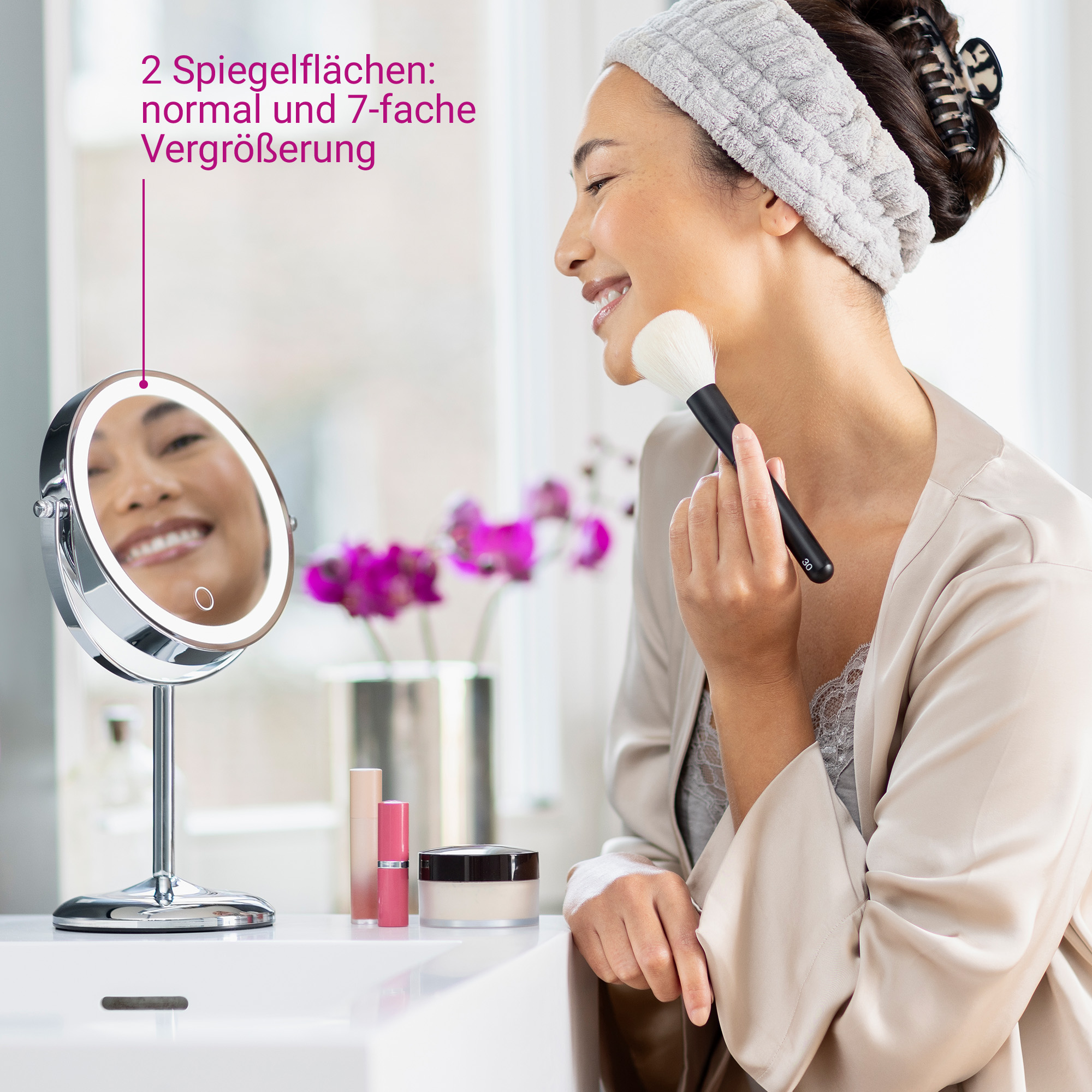 Frau mit Schminkpinsel vor beleuchtetem Spiegel, Make-up-Produkte auf dem Tisch.