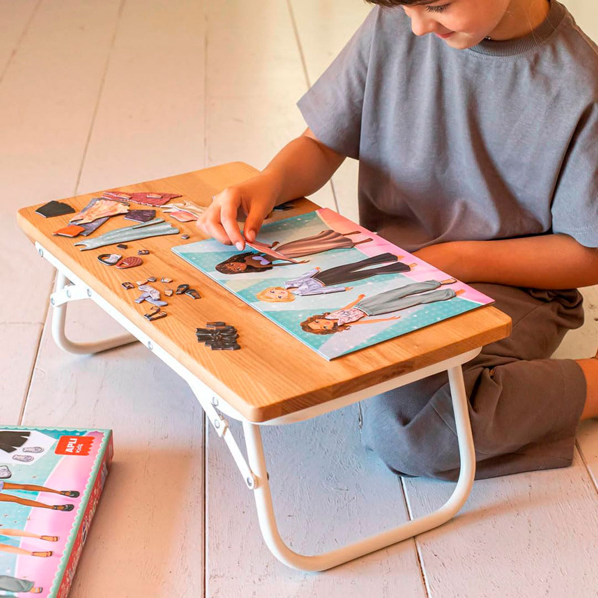 Niño juega con pegatinas en una mesa de madera. Se ven ropa y muñecas.