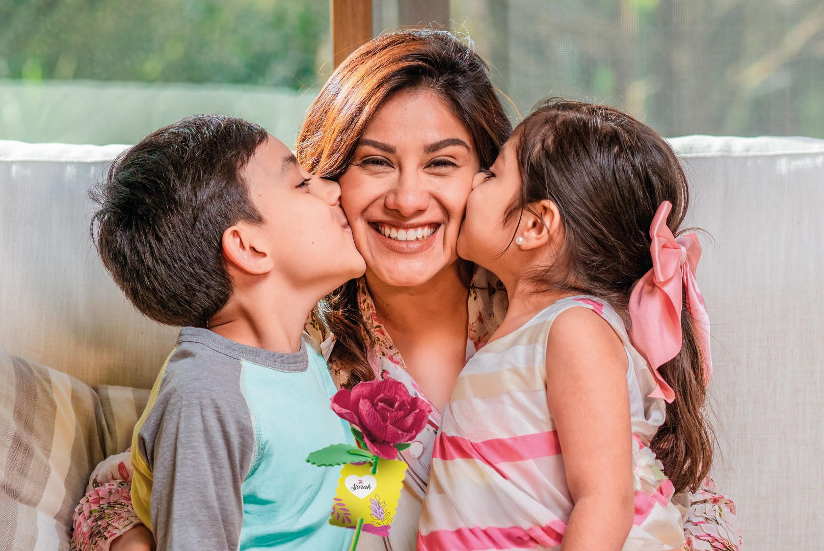 Dos niños besan en las mejillas a una mujer sonriente. Ella sostiene una rosa roja.