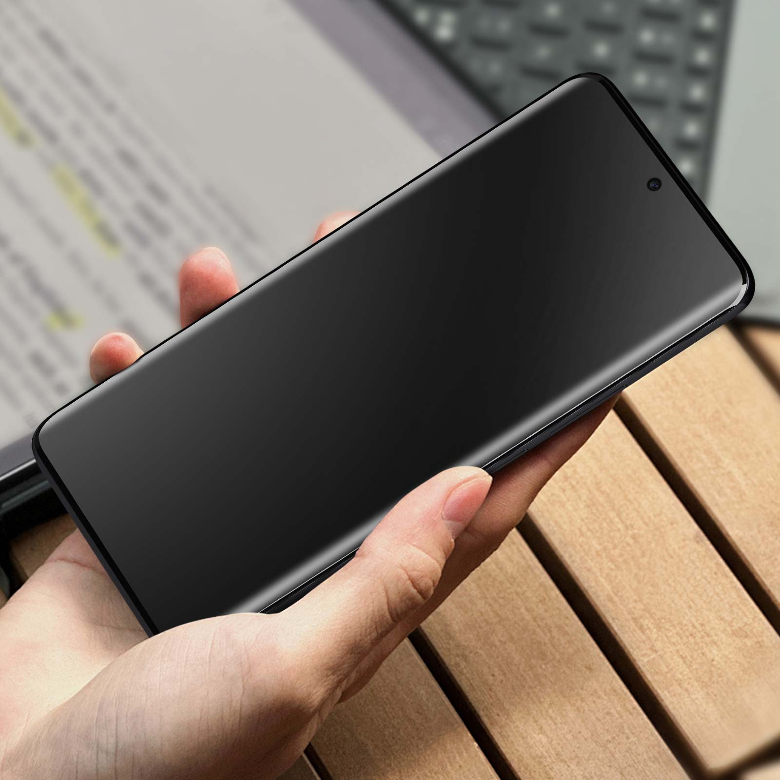 Una mano tiene uno smartphone scuro su uno sfondo sfocato di legno e laptop.