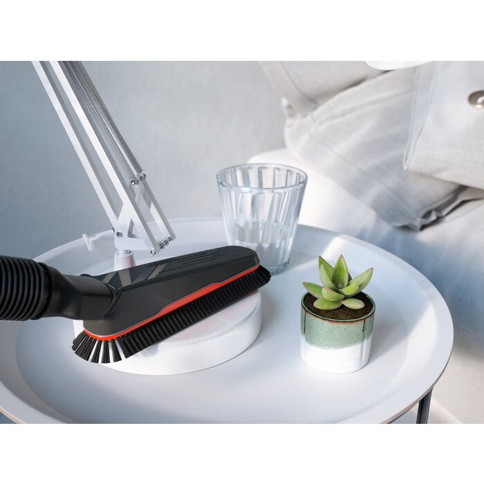 Un accessorio per aspirapolvere con spazzole poggia su un tavolo bianco accanto a un bicchiere e una pianta.