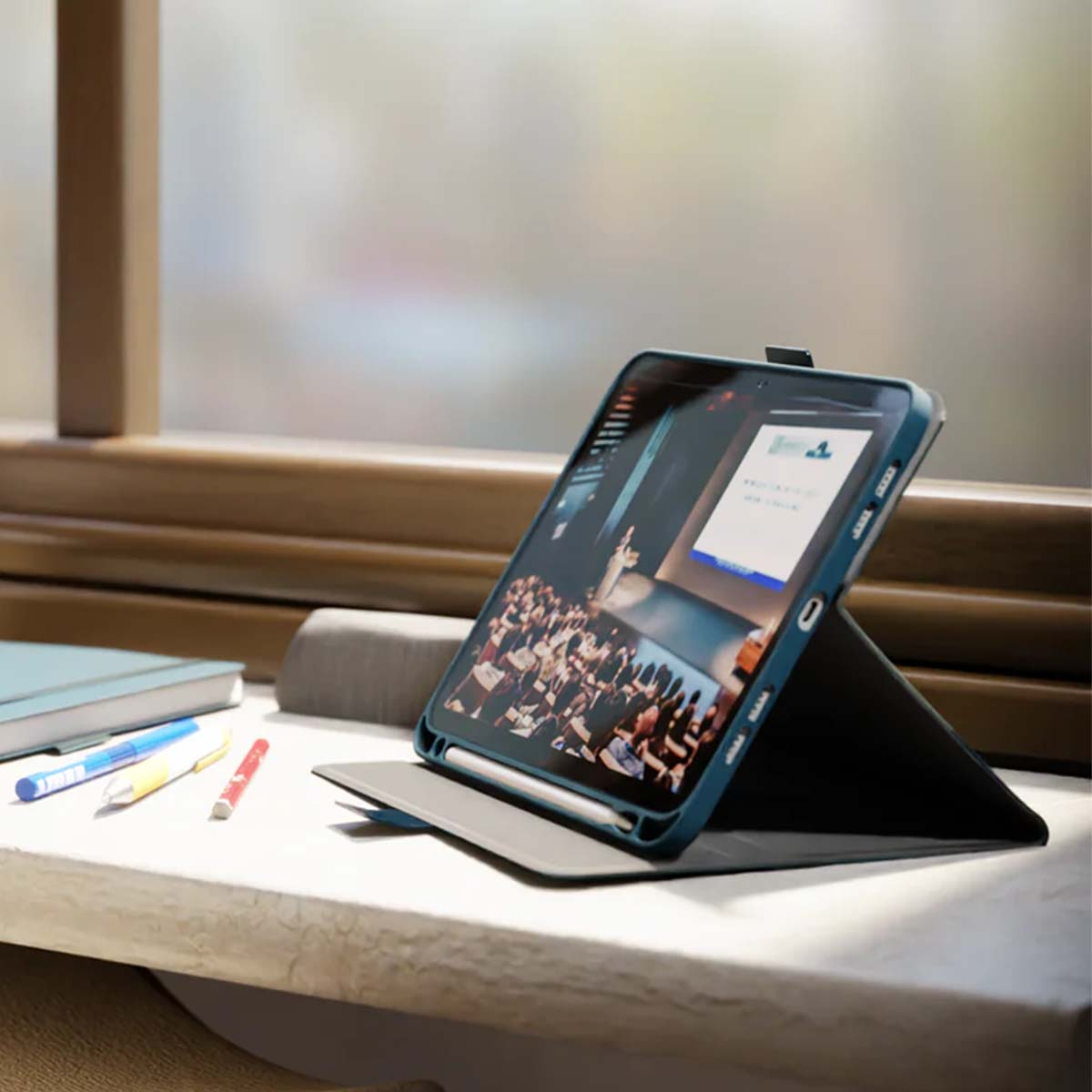 Un tablet che mostra una presentazione poggia su una scrivania accanto a penne e un taccuino.