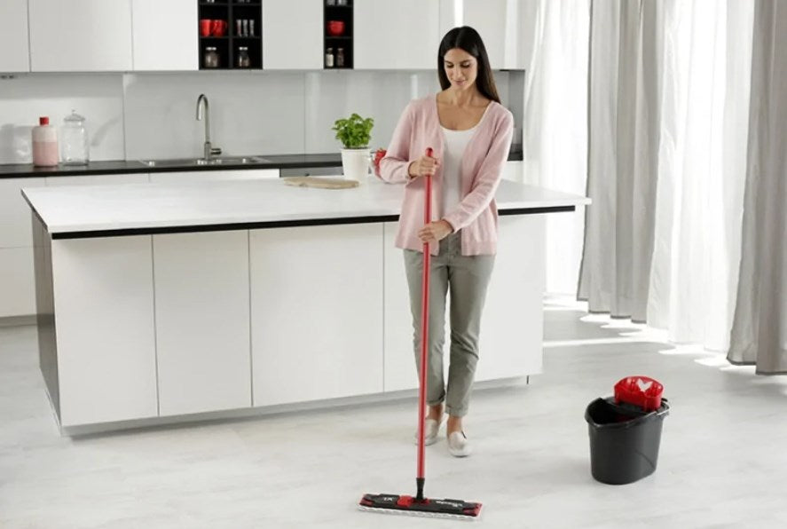 Una donna pulisce un pavimento luminoso della cucina con uno spazzolone rosso.