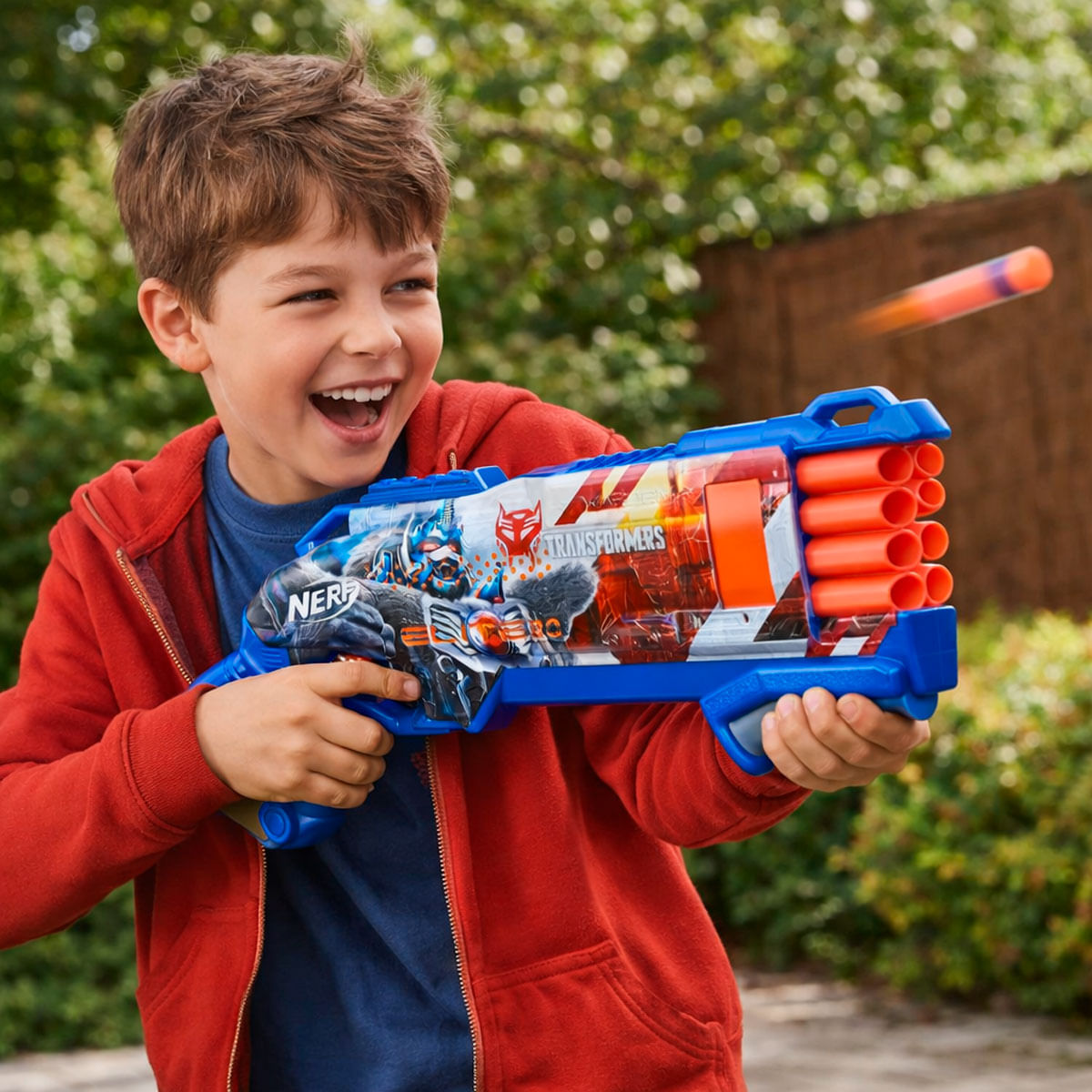 Un niño con sudadera roja ríe mientras sostiene una pistola de juguete Nerf Transformers azul.