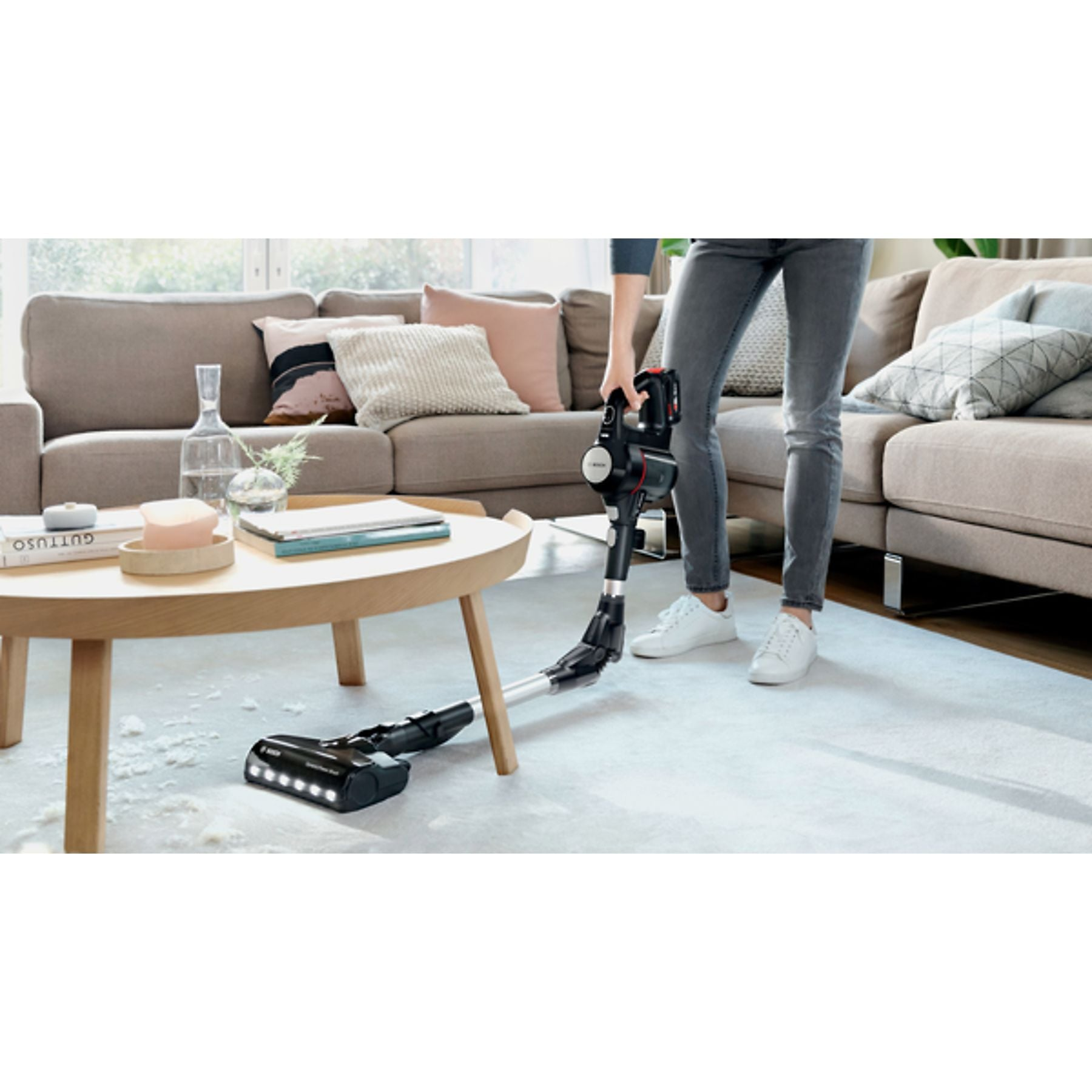 Eine Person saugt einen hellen Teppich mit einem schwarzen Akku-Staubsauger in einem Wohnzimmer.