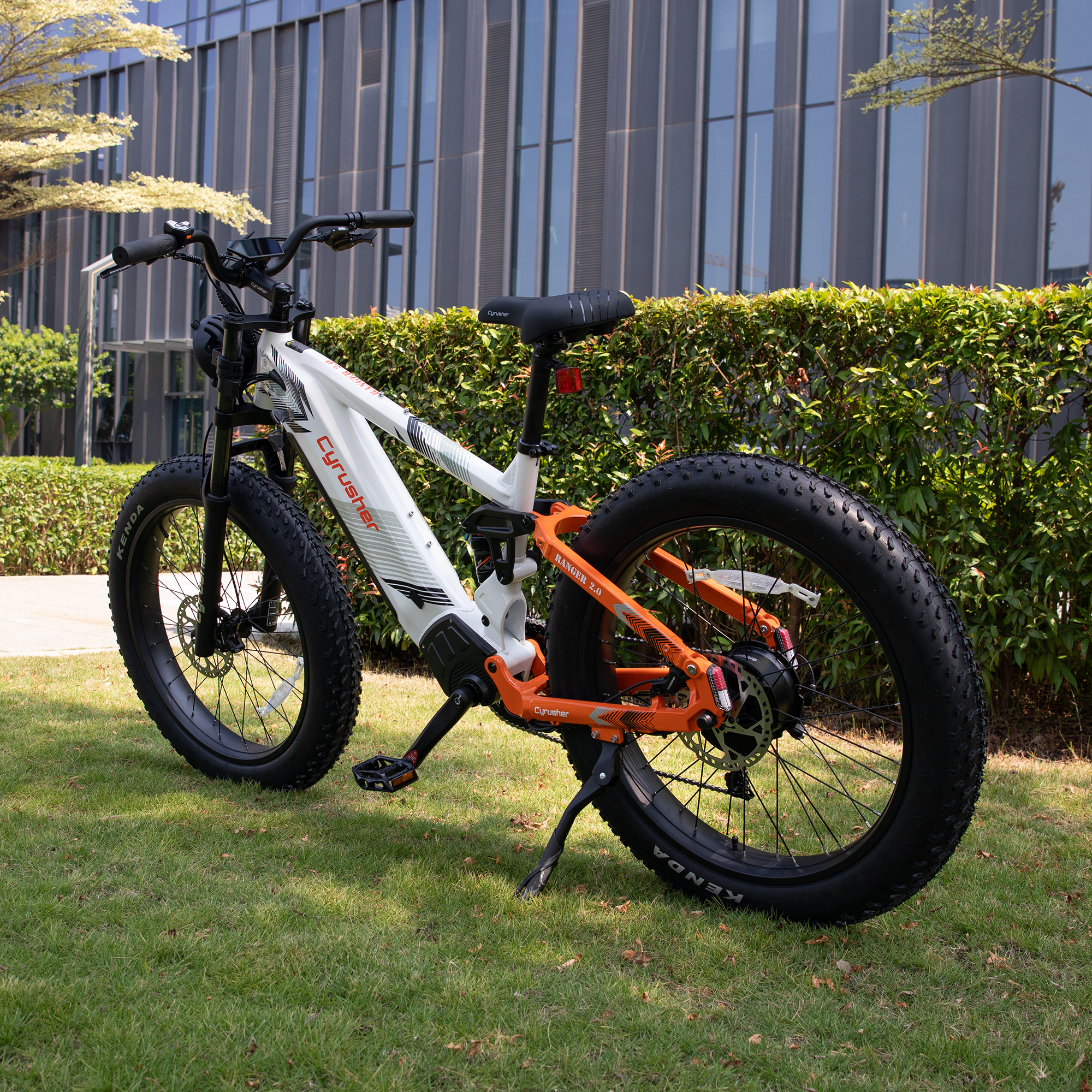 Biało-pomarańczowy rower elektryczny fat bike stoi na zielonej trawie przed nowoczesnym budynkiem.