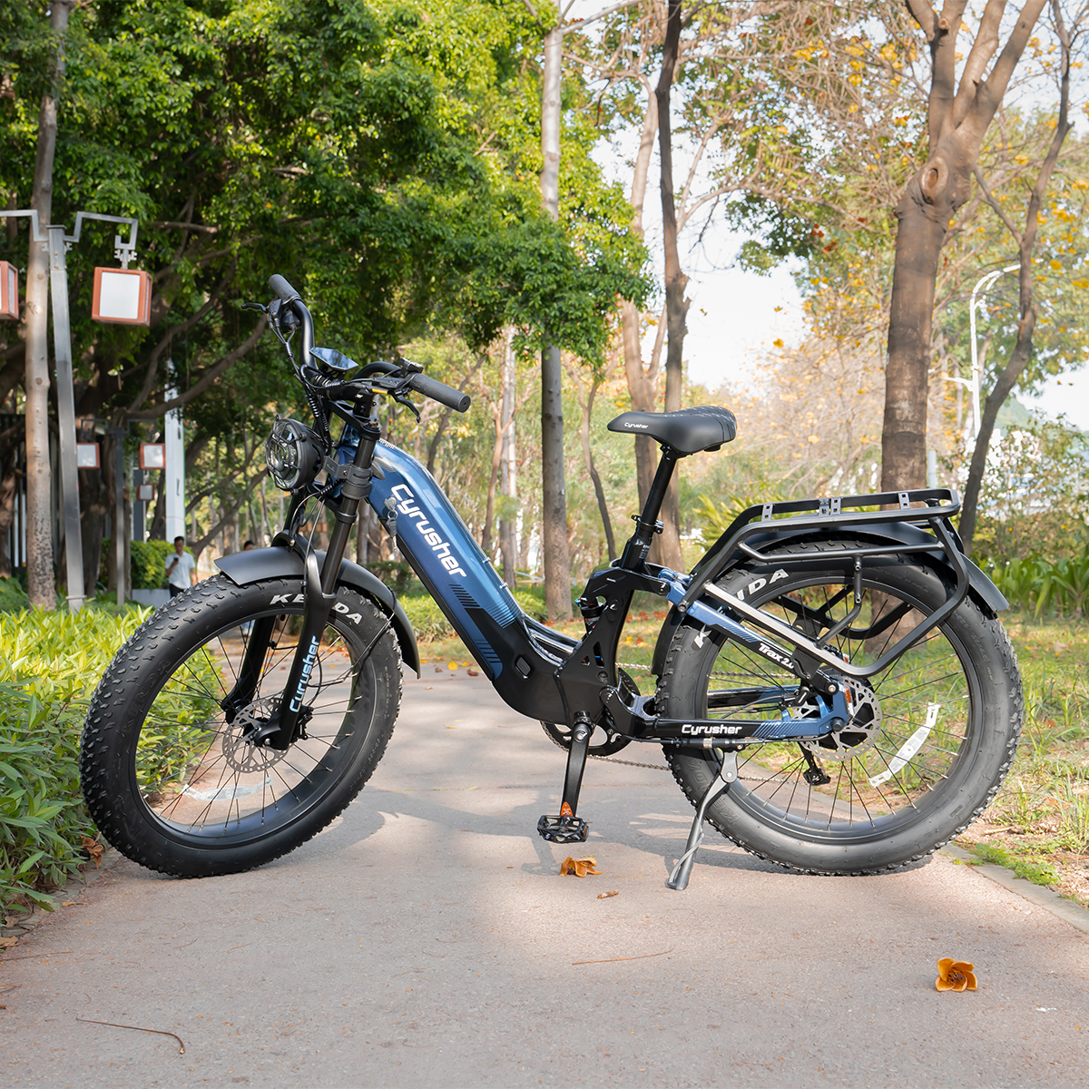 Niebieski rower elektryczny fatbike z bagażnikiem zaparkowany na wybrukowanej ścieżce w parku.