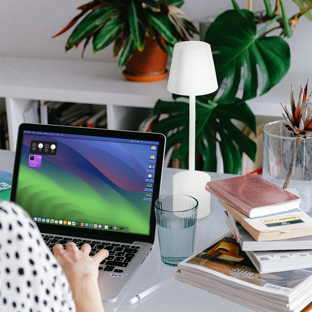 Un laptop con sfondo blu-verde, accanto una lampada, un bicchiere d'acqua e libri.