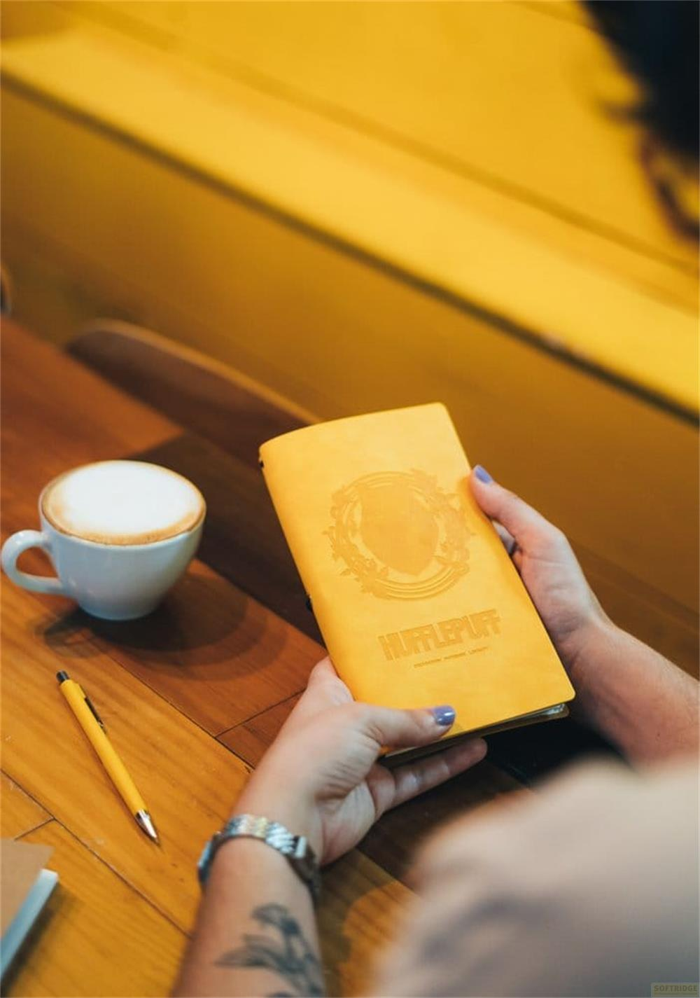 Una taza de café amarilla junto a un cuaderno amarillo con el relieve de Hufflepuff.