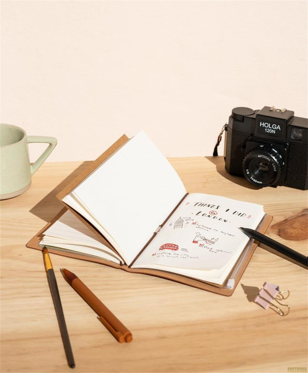 Un diario de color canela con dibujos y texto abierto sobre un escritorio de madera. Una cámara y una taza están cerca.