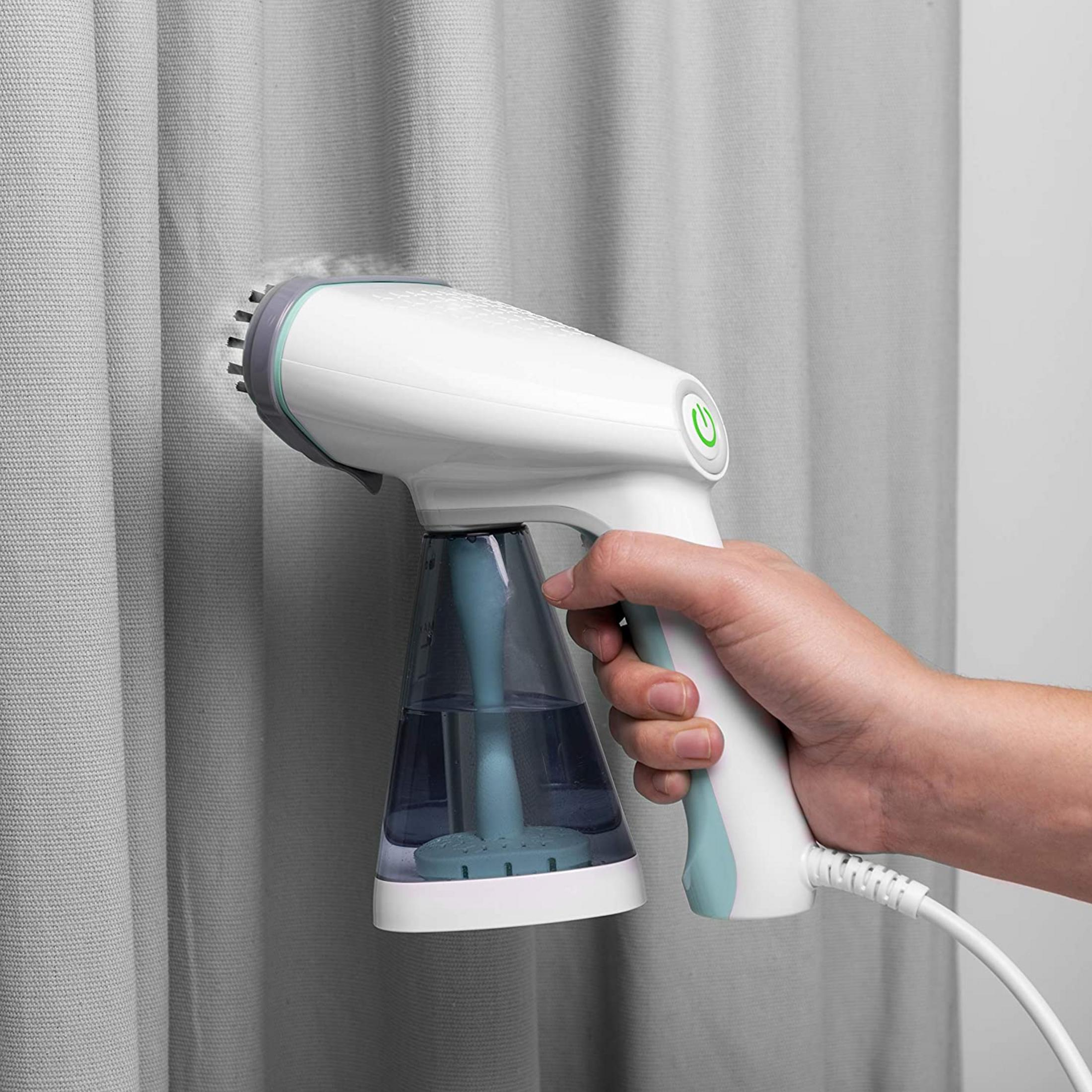 Una mano sujeta un vaporizador de tela blanco que alisa cortinas grises.