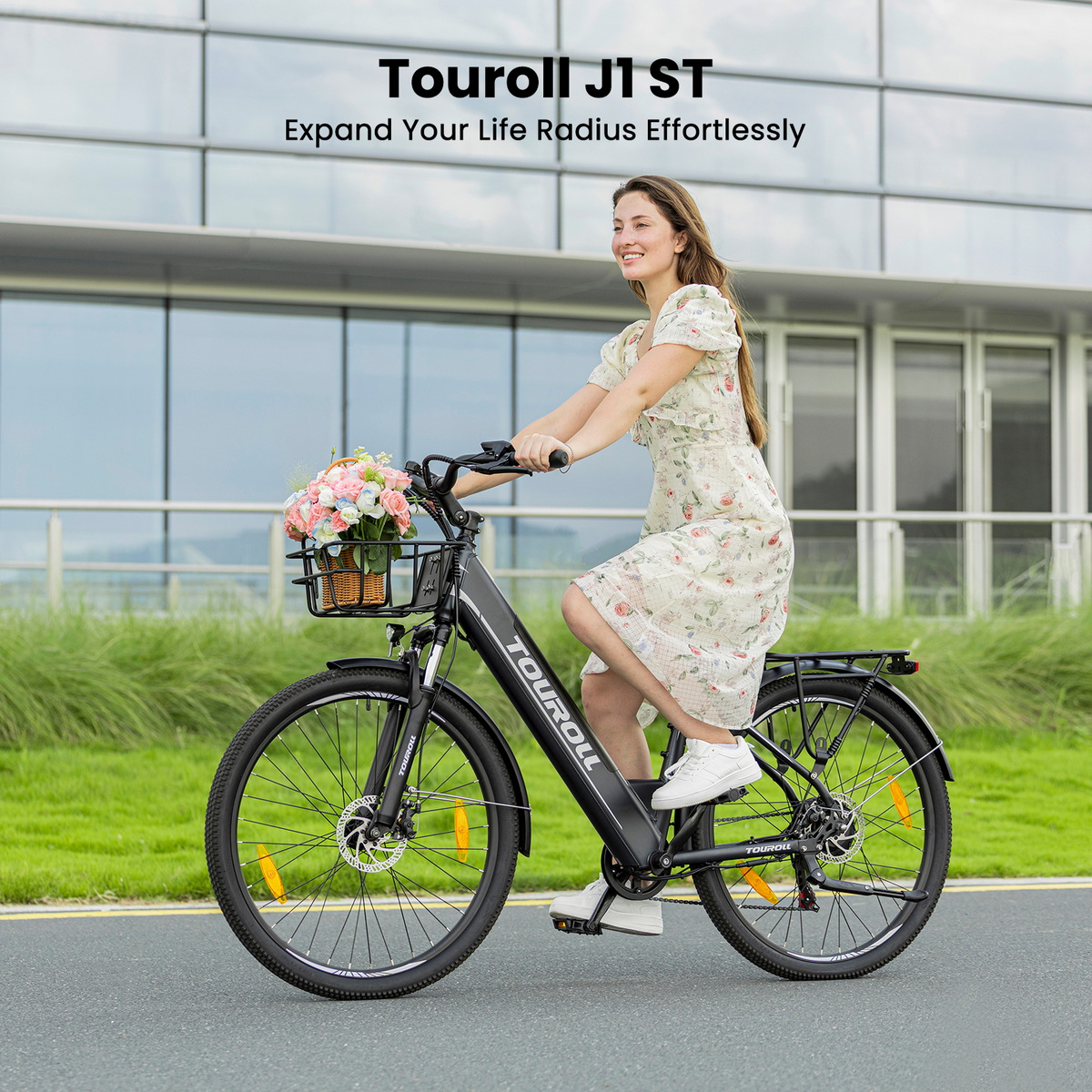 Frau im geblümten Kleid fährt schwarzes E-Bike mit Blumenkorb an modernem Gebäude vorbei.