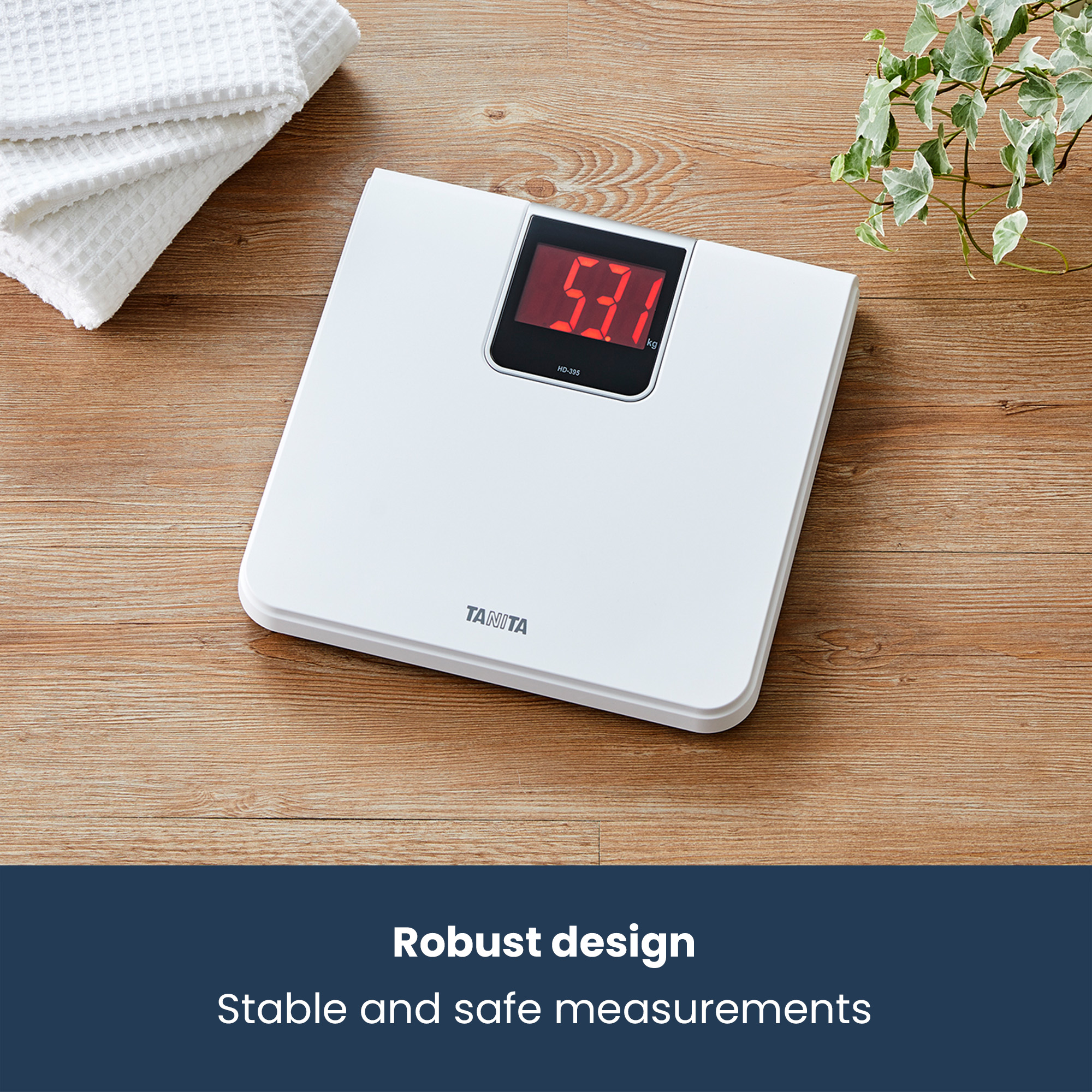 White digital scale showing 53.1 kg on a wooden surface with towels and plant.