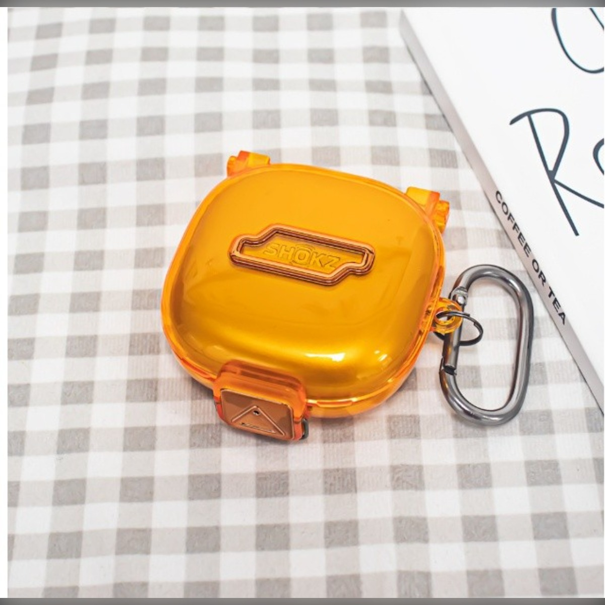 Orange SHOKZ Hülle mit Karabiner auf kariertem Hintergrund.