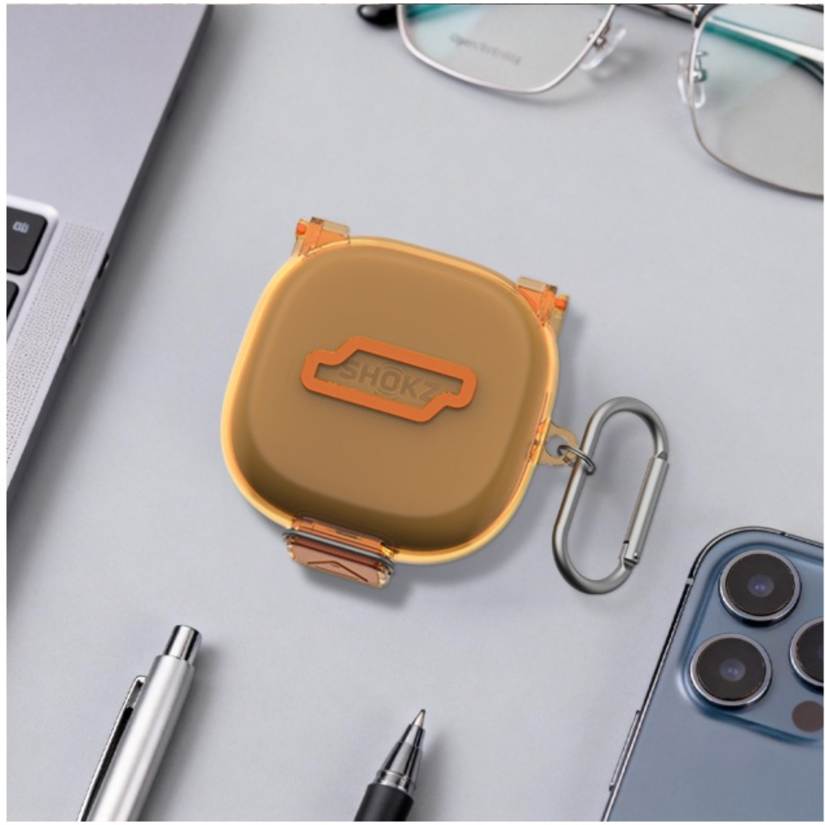 Ein bernsteinfarbener Kopfhörerbehälter mit dem SHOKZ-Logo, neben einem Laptop, Brille, Stiften und Smartphone.