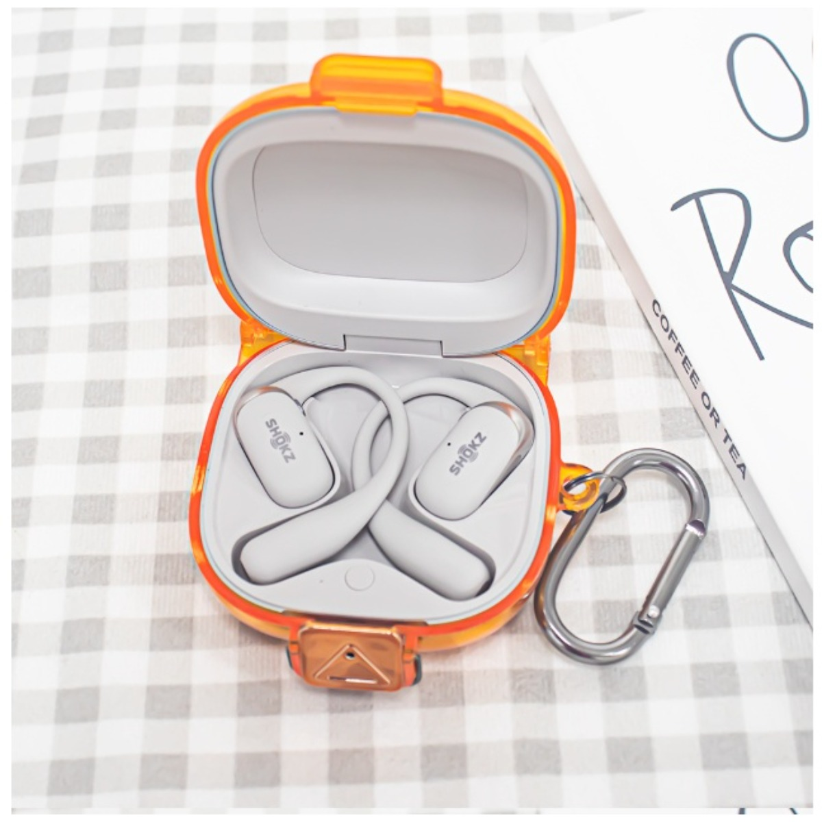 Graue Kopfhörer mit Bügel im geöffneten orangefarbenen Ladeetui, daneben ein Karabiner.