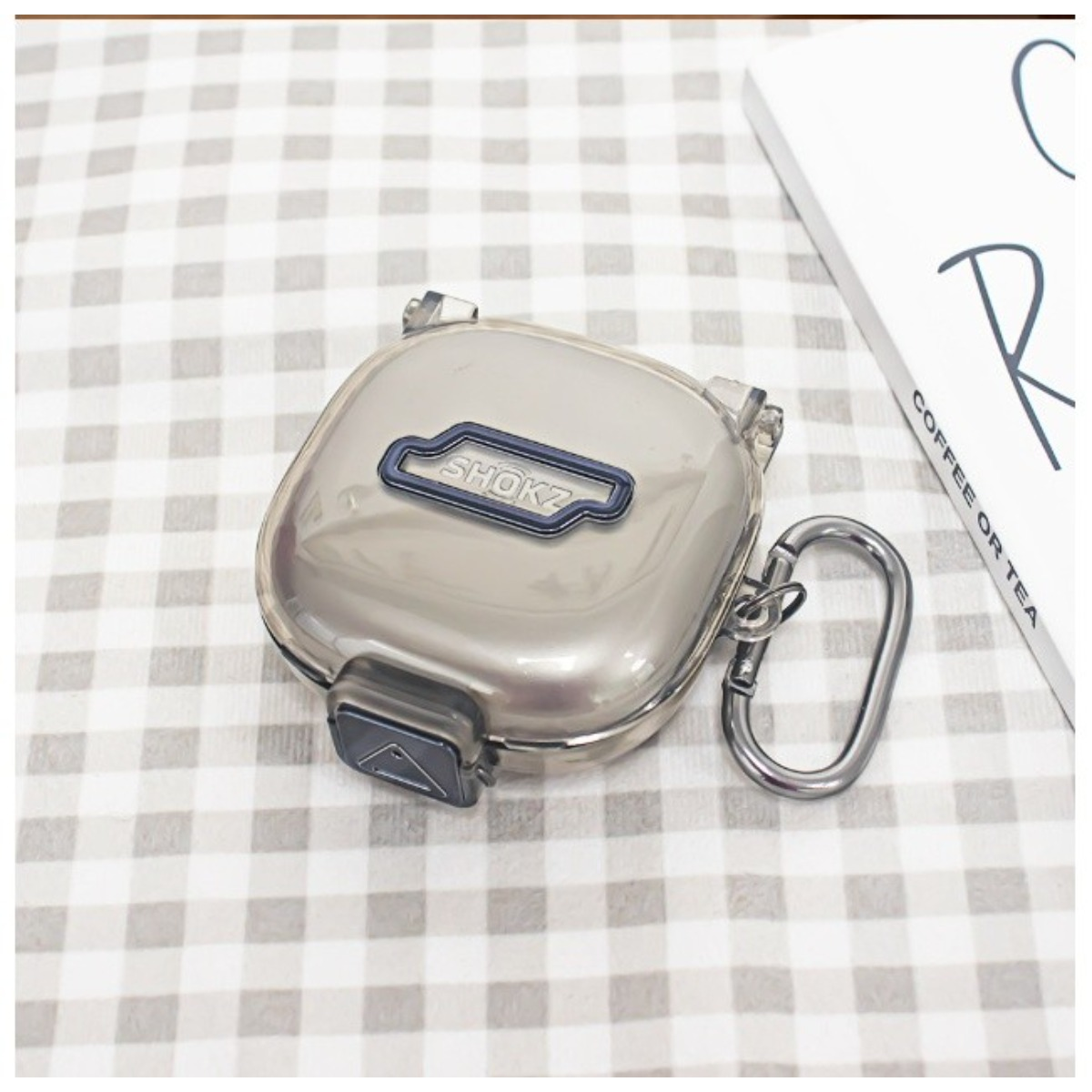 Ein transparenter SHOKZ Kopfhörer-Etui mit Karabiner auf kariertem Hintergrund.