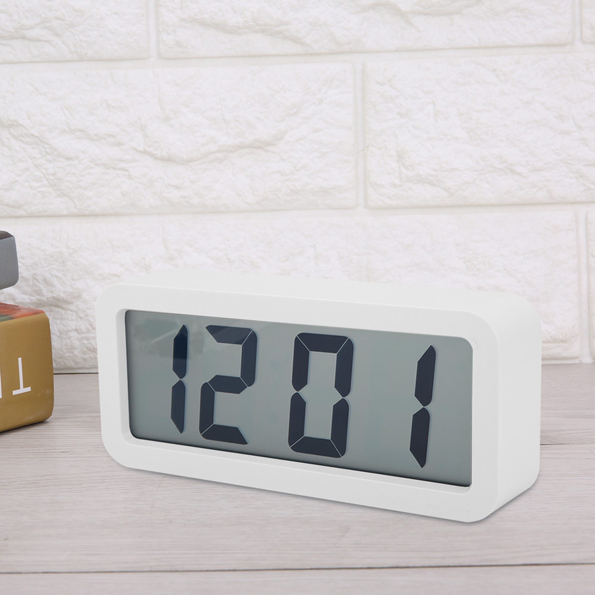 Ein weißer digitaler Wecker zeigt 12:01 auf einem Holztisch vor einer Ziegelwand.