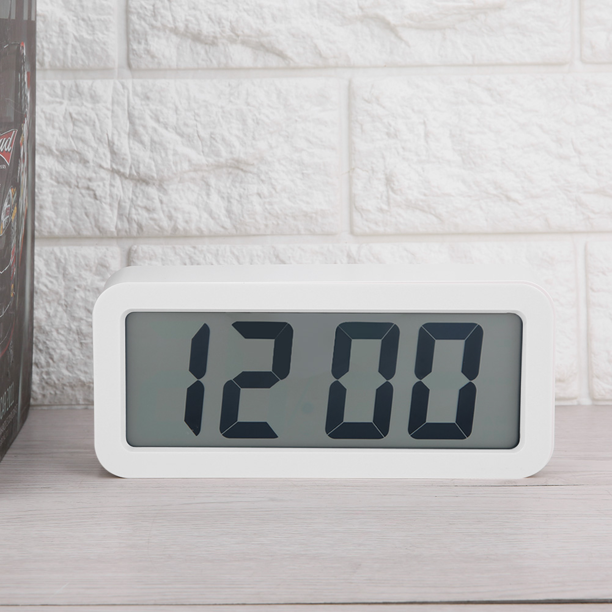 Eine weiße Digitaluhr zeigt 12:00 auf einer hellen Holzoberfläche an. Weiße Ziegelwand im Hintergrund.