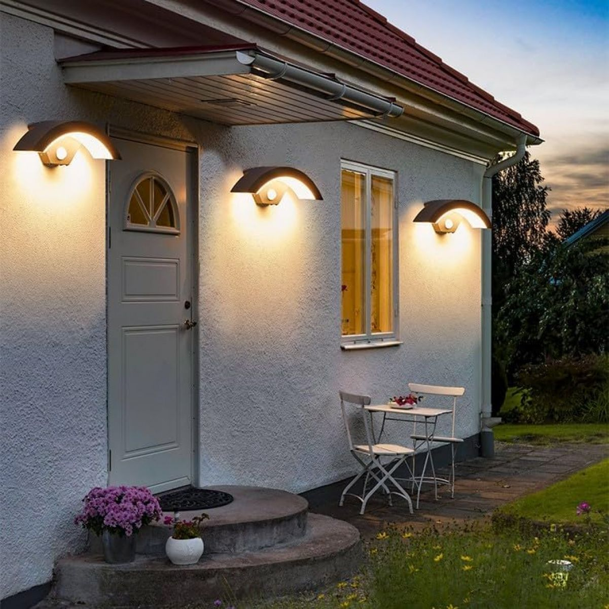 Hauswand mit drei Wandleuchten, Tür, Fenster, Tisch und Stühlen im Abendlicht.