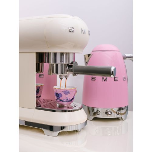 Een witte espressomachine zet koffie in een kopje met patroon, naast een roze Smeg waterkoker.