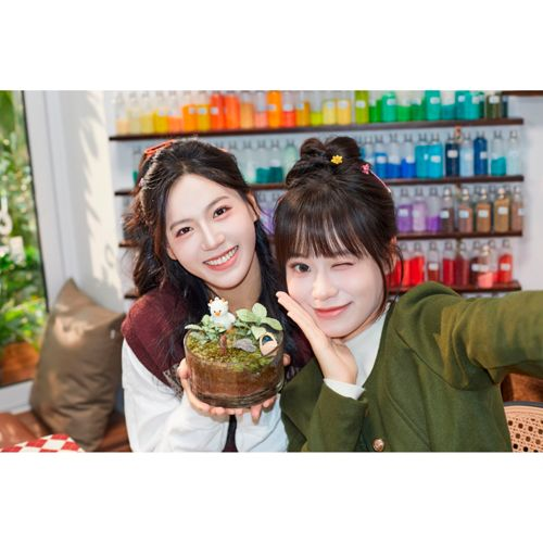 Twee lachende vrouwen poseren met een klein terrarium. Achter hen planken met kleurrijke flessen.