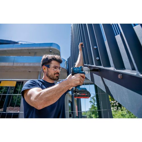 Man met veiligheidsbril en oordopjes boort metalen hek met blauwe boormachine.