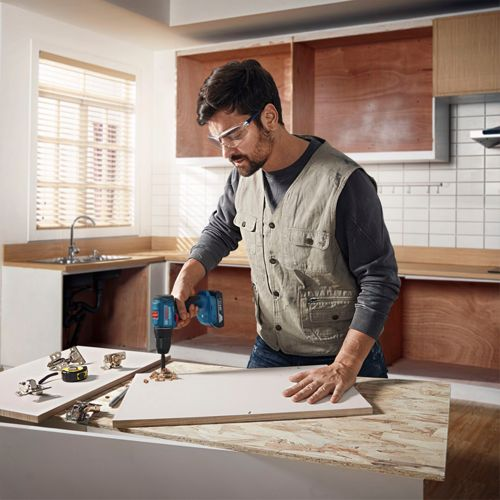 Uomo fora foro in pannello bianco, attrezzi vicini. Cucina sullo sfondo.