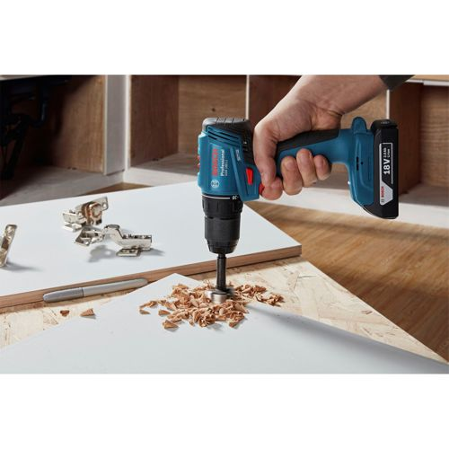 Una mano tiene un trapano Bosch blu che fa un foro in una tavola bianca con trucioli.