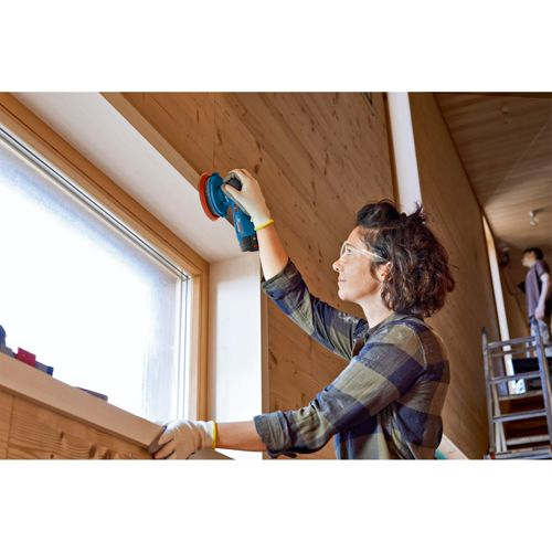 Eine Frau schleift eine Holzwand mit einem blauen Exzenterschleifer neben einem Fenster.