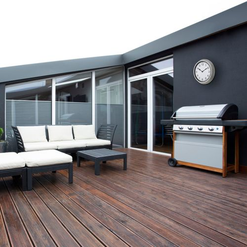 Eine Terrasse mit Sofa, Tisch und Grill. Holzdielenboden und dunkle Wände.