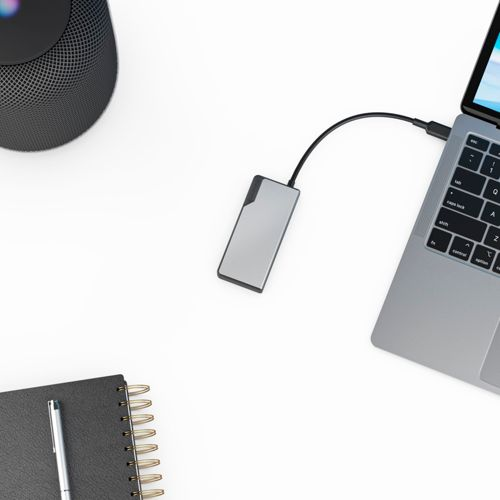 Un hard disk esterno argentato è collegato a un laptop argentato su una scrivania bianca. Nelle vicinanze si trovano un altoparlante nero e un taccuino.