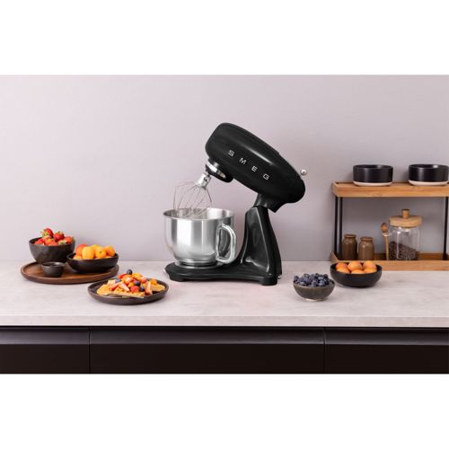 Una batidora amasadora SMEG negra con bol de acero inoxidable sobre encimera con fruta y huevos.