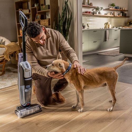 Uomo inginocchiato vicino a cane e aspirapolvere in stanza luminosa con cucina.