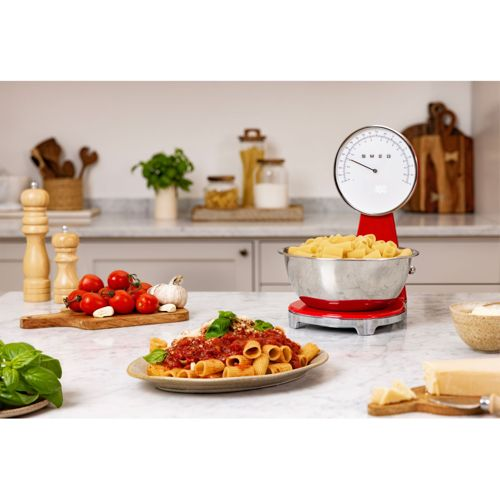 Balanza de cocina roja con pasta, plato de rigatoni, tomates, ajo, albahaca y queso.