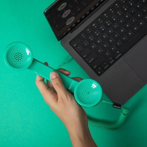 Una mano tiene una cornetta telefonica retrò turchese collegata a un laptop su sfondo turchese.
