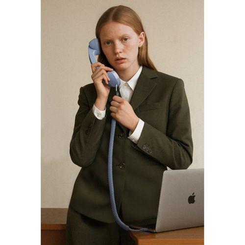 Una donna in tailleur verde scuro tiene un telefono con filo blu, guarda la telecamera.