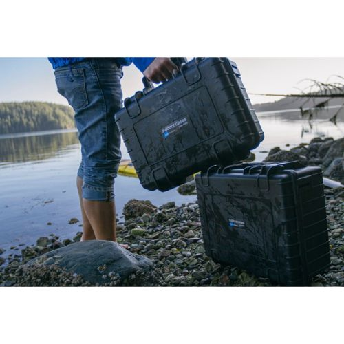 Eine Person in Jeansshorts hält eine schwarze wasserdichte Kiste an einem See. Eine weitere Kiste steht am Ufer.
