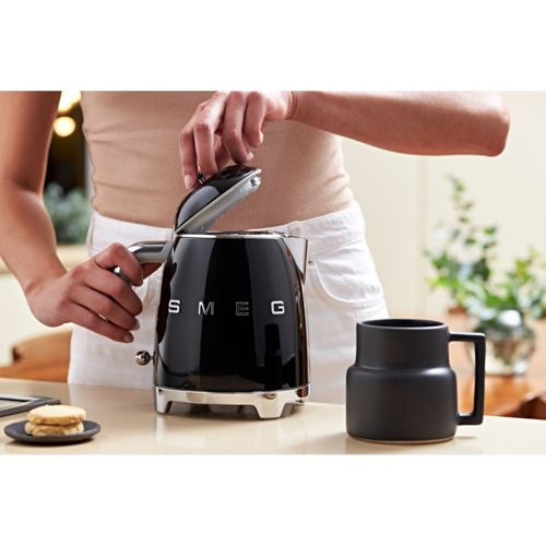 Una persona abre un hervidor Smeg negro junto a una taza negra. Dos galletas.