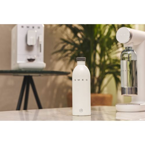Weiße Flasche mit SMEG-Logo steht auf einem Tisch neben einem Kaffeeautomaten.