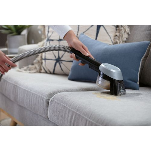Un dispositivo portatile pulisce una macchia su un divano grigio con cuscini blu sullo sfondo.