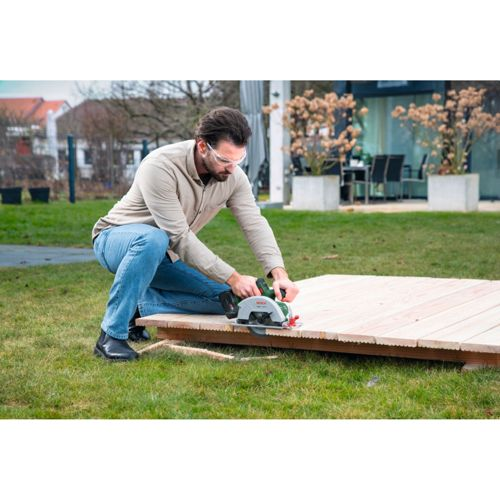 Mann mit Schutzbrille schneidet Holzbrett mit Kreissäge im Freien.