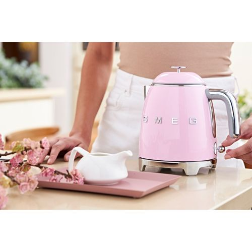 Een roze Smeg waterkoker staat op een aanrecht naast een witte melkkan en bloemen.