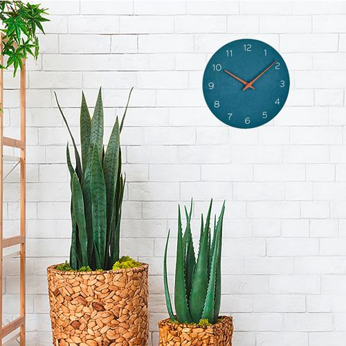 Eine blaue Uhr mit orangefarbenen Zeigern hängt an einer weißen Backsteinwand neben zwei großen Topfpflanzen.