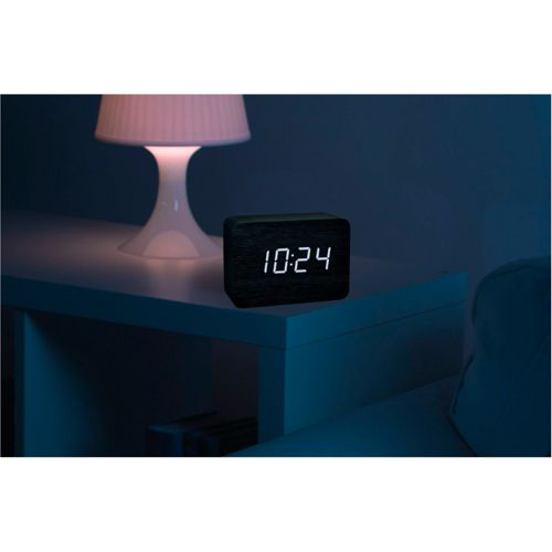 Ein digitaler Wecker zeigt 10:24 auf einem Nachttisch neben einer Lampe.