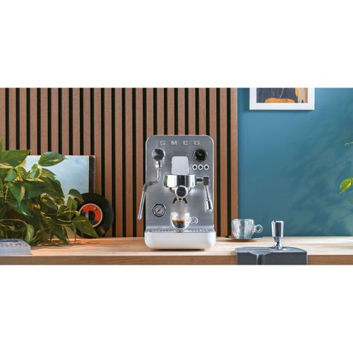 Una cafetera espresso Smeg plateada con un pequeño espresso, junto a una planta.
