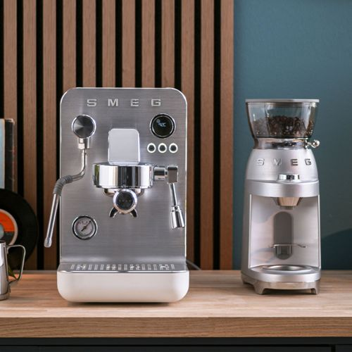 Máquina de espresso y molinillo de café SMEG plateados sobre superficie de madera.